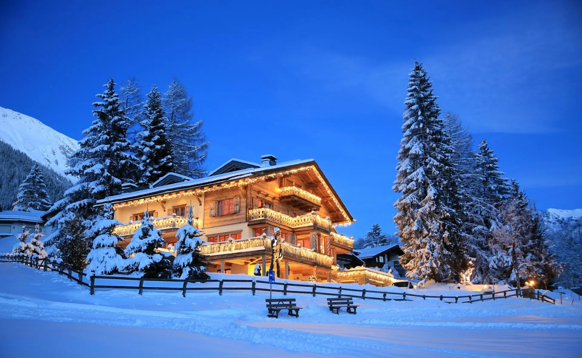 Chalet Kalem in Klosters lit up at dusk in deep snow, framed by fir trees