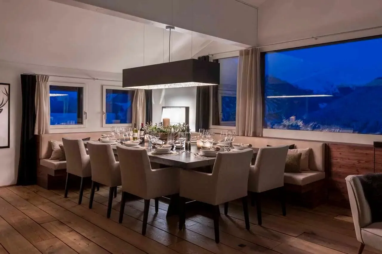A dining table for ten in front of panoramic windows at dusk, Chalet Kyla, Davos